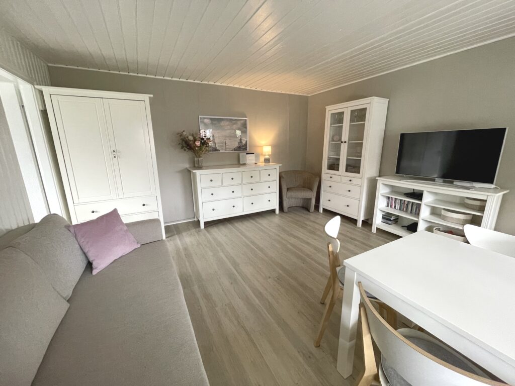 Wohnzimmer, Blick auf die Wand gegenüber vom Fenster. An der Wand steht eingroßer weißer Schrank und eine weiße Kommode.