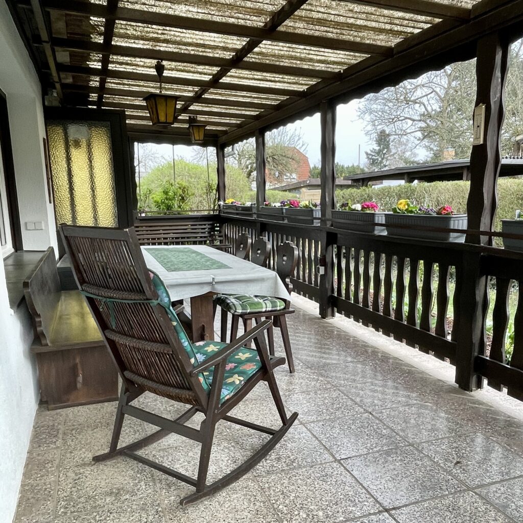 Blick auf die Terasse, Überdachung mit dunklem Holz, Holzgeländer. Im Vordergrund ein hölzerner Schaukelstuhl vor dem großen Tisch.